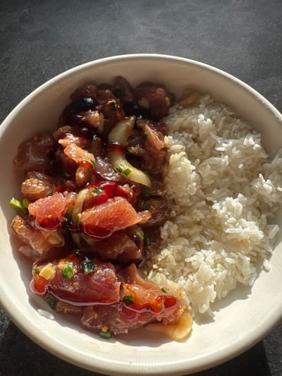 A light-colored, off-white bowl is depicted from a top-down perspective, resting on a dark, textured surface under direct natural lighting. The bowl contains a meal composed of two distinct sections. The left half is filled with poke, which consists of cubed raw fish, likely tuna, coated in a glossy, reddish-brown sauce. This poke mixture includes visible slices of white onion and chopped green onions. The right half of the bowl is occupied by plain white rice, with individual grains clearly visible. There are no people, actions, or interactions depicted. This scene is located in East Honolulu, United States.