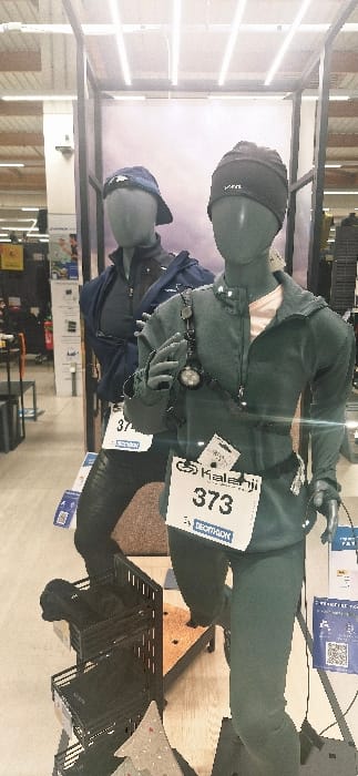 Two mannequins stand in a retail display case. The top mannequin is dressed in a dark green outfit with a headlamp and gloves. A race number, "373", is pinned to the jacket. The lower mannequin wears a navy blue jacket and a cap. The case is illuminated by overhead lights. Various retail shelves and merchandise are visible in the background.