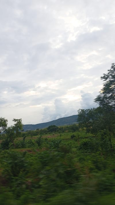 The image depicts a landscape with rolling hills under a cloudy sky. In the foreground, dense green foliage is blurred, suggesting motion or a close-up perspective. Further back, trees and vegetation cover the hillsides, leading up to a more distant, shadowed mountain range. The sky is predominantly filled with soft, diffused light filtering through the clouds, with some brighter patches indicating the sun's presence. The scene appears to be a natural, undeveloped area, characteristic of the rural outskirts of Bokana, Nigeria. There are no people or human structures visible in the frame.