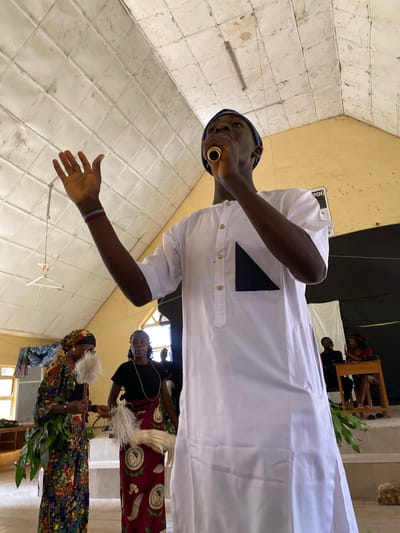 In Dutsen Kato, Nigeria, three individuals are captured in a photograph, with the perspective rotated by 90 degrees clockwise. The primary subject is a person in the foreground, dressed in a white, long-sleeved garment with gold buttons and a dark blue geometric accent on the left chest. They wear a dark blue cap and a gold ring in their lower lip. Their left arm is extended, palm open, and they appear to be speaking or singing into a golden object held near their mouth.
Behind this central figure, two women are visible. The woman on the left is wearing a black top and a patterned skirt featuring red and white motifs. She is looking towards the viewer. The woman on the right is dressed in a colorful, patterned attire, including a headscarf, and holds a whisk-like object in her right hand. She is also holding what appears to be greenery or branches in her left hand.
The setting seems to be indoors, with tiled walls and a yellow ceiling. In the background, a white cloth is draped, and a wooden chair with a person seated on it is partially visible. There is also a sign with some text, but it is mostly obscured. The overall lighting suggests daytime. The expressions of the people are difficult to ascertain with certainty due to the image rotation and resolution, but the scene appears to be part of a cultural gathering or ceremony.