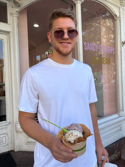A young adult male, with light-colored hair and a light beard, stands outdoors in bright daylight, directly facing the camera with a smile. He wears a white short-sleeved t-shirt and dark-lensed sunglasses with thin metal frames. In his left hand, he holds a paper cup containing a waffle cone filled with two scoops of ice cream (one light-colored with visible inclusions, and one darker, purplish scoop), accompanied by a green plastic spoon.
The background identifies the location as Lambertville, United States. Specifically, the male is positioned in front of an ice cream shop named "Sandy's Scoops Ice Cream," with the name displayed in purple and yellow lettering on the shop's large front window. The shop's interior, visible through the window, features a light pink color scheme. White, ornate architectural trim frames the shop's windows and a adjacent white paneled door to the left of the subject. A faint reflection of trees is visible in the window, and a small Coca-Cola sign can be seen inside the shop through the left window pane.