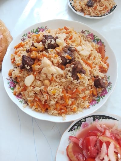 A plate of plov, a traditional Uzbek dish, is centered in the frame, filling most of the image. The plov consists of rice, shredded carrots, chickpeas, and pieces of meat, garnished with cloves of garlic. To the right and slightly behind it, a smaller plate of sliced tomatoes and onions is visible. To the left, a piece of bread, likely lavash, rests on the table. The meal is served on a white, floral-patterned plate. The food is placed on a white table with faint blue lines. The scene appears to be an indoor setting, possibly a home or restaurant, in Jizzakh, Uzbekistan. The lighting suggests it is daytime, with sunlight casting shadows.