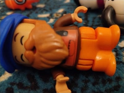 A small toy figure with a blue hat and beard lies on a textured surface. The figure has a tan-colored head with curved lines resembling eyes and a small hole for a mouth. It wears a brown shirt and bright orange pants and boots. One hand is raised in a curved "C" shape. Other toy figures are partially visible in the background.