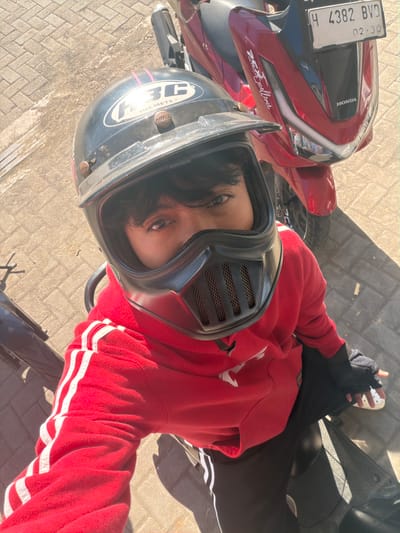 A person wearing a black helmet and a red hoodie sits on a motorcycle. The helmet has a black mask with a grid pattern. The hoodie has white stripes on the sleeves. The motorcycle is red and has a license plate that reads "H 4382 BVD 02.30". The ground is paved with bricks. The scene appears to be in Jipang, Indonesia.