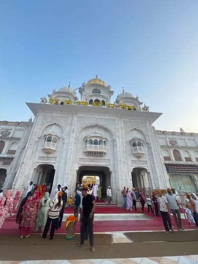 A low-angle, eye-level outdoor shot captures the ornate white marble facade of the Golden Temple complex in Amritsar, India, under a clear blue sky. The main structure features multiple domes and intricate carvings. Yellow lettering is prominently displayed above the central entrance arch. A crowd of people, dressed in a variety of colorful clothing, including traditional Indian attire and western wear, are seen walking on red carpeted steps leading into the temple. Some individuals are wearing turbans, indicating a religious or cultural significance. In the foreground, a basket filled with orange fabric sits on the ground, and several people are walking barefoot, suggesting it is a sacred site. The overall scene conveys a sense of bustling activity and religious devotion.