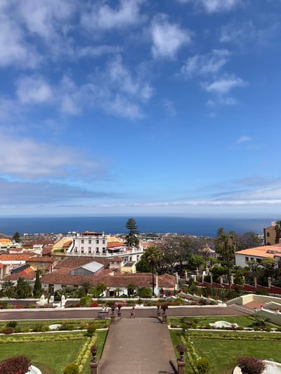 The image shows a panoramic view of La Orotava, Spain, from an elevated vantage point. In the foreground, a well-maintained garden with manicured lawns and colorful flowerbeds leads to a stone pathway. Two individuals are visible on the pathway, one appearing to walk and the other standing.

Beyond the garden, the scene transitions to a town with red-tiled roofs and white buildings, characteristic of Spanish architecture. A large white building with multiple windows and balconies stands out prominently. Further in the distance, the deep blue of the Atlantic Ocean stretches to the horizon, meeting a bright blue sky dotted with scattered white clouds. The overall impression is one of a picturesque coastal town on a clear, sunny day.