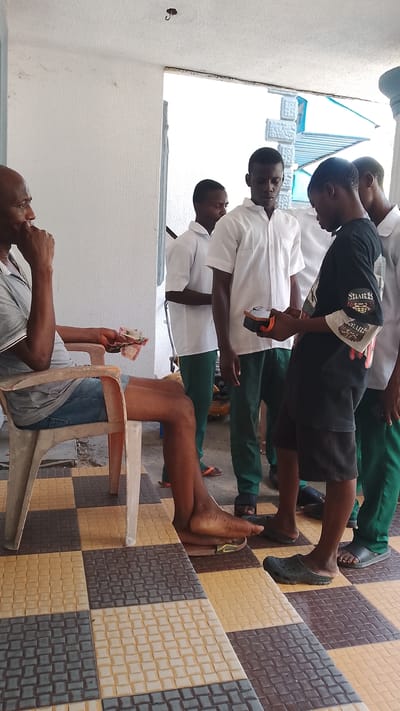 A man is sitting on a plastic chair with money in his hand. He is wearing a gray t-shirt and denim shorts. He is sitting on a tiled porch in what appears to be Asubi, Nigeria, based on the information provided. There are four teenagers standing near him. Three of the teenagers are wearing white shirts and green pants. One of the teenagers is wearing a black t-shirt with a graphic design and black shorts, and is holding a small, dark electronic device. All of the teenagers are wearing sandals.