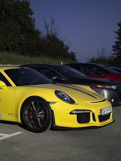 A bright yellow Porsche GT3 is parked on asphalt in front of a line of other cars, including a dark colored sedan and a red vehicle, under a dark blue sky with trees in the background. The yellow Porsche is in the foreground, facing right, with its front wheel visible, along with its license plate which reads "CB 1919PE". The car has a black stripe running down the hood. The environment suggests an outdoor parking area in Arbanassi, Bulgaria, likely during dusk or early evening. No people are visible in the frame.