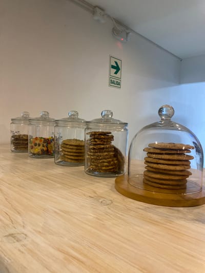 The image shows a wooden counter in a shop in Punta Hermosa, Peru (latitude: -12.33, longitude: -76.827), featuring four glass jars filled with cookies and a glass cloche with a stack of cookies. The jars are arranged in a row, with the cloche to the right. The jars contain various cookies, including chocolate chip, M&M, and plain. The cloche holds a stack of about 10 cookies. The counter is a light wood, and the wall behind is white. A green arrow sign with the word "SALIDA" (Spanish for "exit") is on the wall. The counter is empty, suggesting that the shop is closed. The scene suggests that the shop may be a bakery or cafe.
