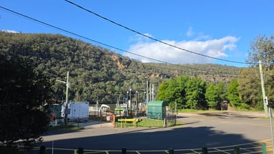 This is an eye-level, wide shot of a ferry landing area in Wisemans Ferry, Australia. The scene is set against a backdrop of a steep, densely forested hill with rocky outcrops. The sky is clear blue with scattered white clouds.

In the foreground, a paved road with yellow markings leads towards the ferry landing. There are some dark green bollards and a yellow barrier across the road, indicating a closed section.

The ferry landing itself is a series of structures and equipment. There's a white trailer-like building on the left, a dark green covered structure in the center, and various poles with lights and what appear to be communication antennas. A black metal fence encloses parts of the landing area. Signs are visible, including one with a red circle and diagonal line over a person symbol, and a yellow "ROAD CLOSED" sign.

A calm body of water, likely the Hawkesbury River, is visible behind the ferry landing structures. The opposite bank of the river is also densely forested, mirroring the hill in the background. Several mature trees with green foliage are scattered throughout the scene, some closer to the foreground on the right and left. Power lines stretch across the top of the frame. No people are visible in the image.