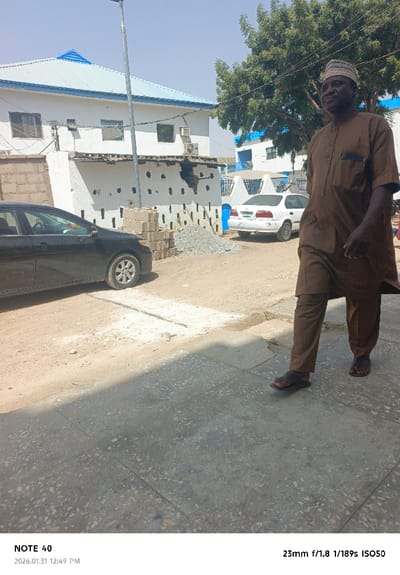 The scene is an outdoor, daytime view in Kofar Ruwa, Nigeria, under clear skies with strong sunlight. In the foreground, a dark-skinned adult male, approximately 50-60 years old, walks towards the left of the frame on a mottled concrete sidewalk. He wears a brown native tunic and trousers, with dark sandals, and a green pen is visible in his left chest pocket. His right leg is slightly raised, indicating mid-stride.
To the immediate left of the man, the concrete sidewalk transitions to an unpaved dirt shoulder. A black sedan, likely a Toyota Corolla, is parked facing left, partially visible with its rear section and driver's side door in the frame. Adjacent to the black car, a stack of approximately 15-20 grey concrete blocks is present, next to a small pile of light-colored gravel.
In the mid-ground, a white sedan, possibly a Honda Civic, is parked further back on the dirt shoulder, facing right. Behind it, a structure with blue accents is partially visible. Dominating the background on the left is a two-story white building with a light blue corrugated metal roof. The lower section of this building features a distinctive wall with multiple rectangular indentations. An external air conditioning unit is mounted on the wall, and multiple rectangular windows are visible on both levels. A tall, slender metal streetlight pole stands between the main building and a large, leafy green tree that occupies the upper right portion of the frame, partially obscuring another building with a blue roof.