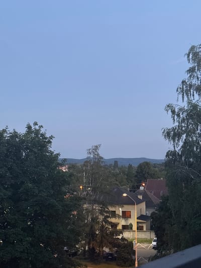 This is a daytime, eye-level shot taken from an elevated vantage point overlooking a residential area in Piešťany, Slovakia. The sky is clear and pale blue, with a faint haze visible towards the horizon. A range of distant, verdant hills forms the backdrop. In the foreground and midground, lush green trees dominate, with dense foliage obscuring much of the view.

Several houses with tiled roofs are visible amongst the trees. One house in the mid-right of the frame is cream-colored with red-tiled roofs and dormer windows. Another, larger house to its left, appears to be tan with darker rooflines and features illuminated windows suggesting interior lighting. A streetlamp with a bright yellow light illuminates a small section of the street and sidewalk in front of this house.

A white car is parked on the side of the road. A dark-colored car is partially visible on the left side of the frame, parked in front of a tree. The scene appears to be a quiet residential street, with no people immediately apparent in the frame. The overall impression is of a peaceful, suburban landscape at dusk or early evening, as indicated by the artificial light from the streetlamp and house windows against the fading daylight.