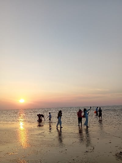 The image displays a coastal scene at sunset in Tritunggal, Indonesia. The sun is visible as a bright orange orb low on the horizon, situated on the left side of the frame, casting a shimmering golden reflection across the calm, shallow water and wet sand extending into the foreground. The sky transitions from a pale blue at the top to hues of orange and pink near the horizon.
Approximately eight to nine individuals are scattered across the wet sand and shallow water. From left to right: two figures are partially submerged, one bent over, possibly digging or observing the water. A small child stands nearby. Further right, a woman in light clothing walks towards the right, carrying an obscured object or child on her front. Adjacent to her, another woman in light attire stands, holding a device (likely a smartphone) aloft, appearing to capture an image or selfie. Behind her, a figure in a dark top and light pants stands. To the far right, two additional figures are partially submerged, one of whom appears to be holding an orange object, possibly a bucket. All individuals are partially silhouetted against the sunset, and their reflections are visible on the wet surface.