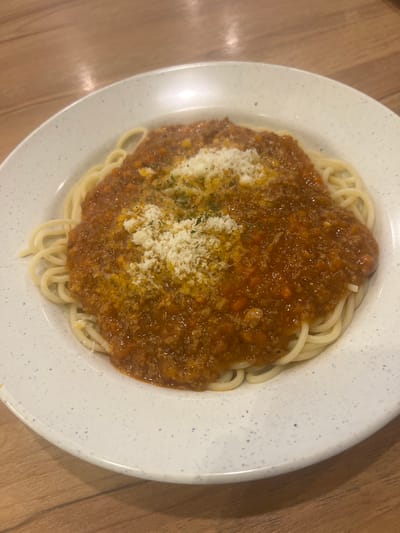 A close-up, overhead shot shows a white plate with a speckled blue rim filled with spaghetti Bolognese. The pasta is thin and golden yellow, topped with a thick, rich, reddish-brown meat sauce. The sauce appears to have finely diced vegetables, possibly carrots and onions, and ground meat. A generous sprinkle of grated white cheese and finely chopped green herbs garnishes the top of the sauce. The plate rests on a wooden surface, likely a table. The lighting is warm and ambient, suggesting an indoor setting. There are no people visible in the frame. The image is sharp and well-lit, focusing entirely on the dish of pasta. The location is identified as Baiyun District, China.