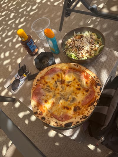 The image depicts an outdoor scene, possibly a patio or garden, with dappled sunlight filtering through an overhead structure, creating a pattern of light and shadow on the ground and objects. The scene is set on a woven textured table.
On the table, a whole pizza with ham and mushrooms is prominently displayed, showing a slightly charred crust. Beside the pizza, a bowl contains a Caesar salad with croutons, shredded cheese, and a drizzle of dressing.
Several other objects are present:
* Two bottles of sunscreen are visible: one dark brown with a yellow cap, labeled "NIVEA SUN Protect & Bronze," SPF 20, and another with a blue body and orange cap.
* A clear plastic cup, partially filled with water, is positioned between the sunscreen bottles.
* A black, octagonal ashtray containing cigarette butts is near the sunscreen bottles.
* A pack of cigarettes and a pair of dark sunglasses are placed next to the ashtray.
* A portion of a backpack is visible in the lower right corner of the image, featuring a zipper.
The overall setting suggests a relaxed, leisurely moment, possibly during a sunny day. The elements present (sunscreen, food, cigarettes) imply an outdoor activity like sunbathing or enjoying a meal outdoors. There is no indication of the city of Delchevo, North Macedonia, or any specific urban features in the image.