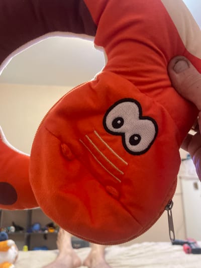 A close-up, high-angle, indoor shot shows a person's hand holding an orange plush toy. The toy is shaped like a cartoonish caterpillar or worm, with large white eyes outlined in black and red. The body of the toy is a bright orange, and it has a zipper on its underside. The background is blurred, but appears to be a bedroom with a bed, shelves with various items, and two bare feet visible in the lower portion of the frame. The lighting suggests it is daytime, with light coming from an unseen window. No information about the city of Laishevo, Russia, can be determined from the image.