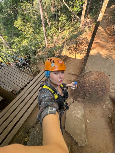 A person wearing an orange helmet and a harness stands on a wooden platform in a forest. They hold a water bottle near a large, rounded rock. The trees form a dense canopy, filtering sunlight. A zipline cable is visible stretching across the scene. The ground is a mix of dirt and stone, with exposed roots. The angle suggests a selfie taken from above.