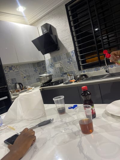 The image displays an indoor kitchen or utility space, viewed from a slightly elevated perspective looking down onto a countertop and back wall. The original image is rotated 90 degrees counter-clockwise.
On the left side of the frame (which would be the top when correctly oriented), there is a window secured with dark black bars. Below the window is a sink area with a silver faucet, from which a light brown plastic bottle with a red cap/spout appears to be hanging.
The middle ground features a wall adorned with distinctive blue and white patterned tiles, arranged in a grid of various geometric and abstract designs. A counter runs along this tiled wall, holding several kitchen items, including a silver kettle and other utensils. Further to the left, sleek black and white cabinets or appliances are visible.
The right side of the image (which would be the bottom when correctly oriented) is dominated by a white countertop, possibly with a subtle marble pattern. Various items are arranged on this surface:
* A clear plastic cup contains a dark brown liquid, likely a beverage.
* Adjacent to it is a dark-colored bottle with a red cap, featuring "BB" written in white on its label.
* Another empty clear plastic cup and a loose blue plastic bottle cap are also present.
* A roll of white paper towels sits prominently on the counter.
* A silver knife with a dark handle is placed next to the paper towels.
* A human hand, with a medium skin tone, is visible holding a dark-colored smartphone.
* A white plate is partially visible at the top edge of the countertop.
The overall lighting suggests an interior setting, possibly under artificial light, with bright reflections on the polished countertop and a visible light fixture on the ceiling or wall in the lower-left area of the frame (when correctly oriented). The scene captures a casual moment in the kitchen, perhaps during the preparation of drinks or a break, with someone engaging with their smartphone.