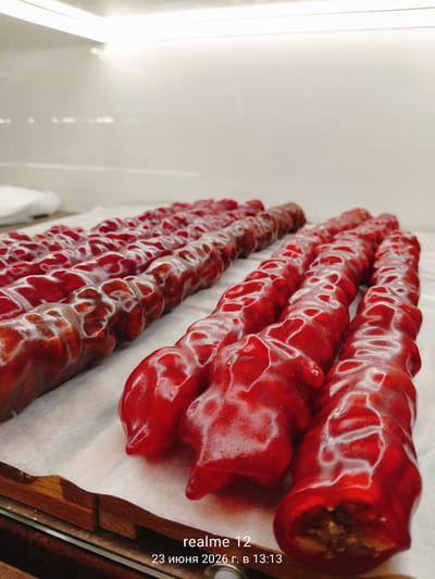 This image shows a close-up, eye-level view of multiple strings of churchkhela, a traditional Georgian candy made from grape juice and nuts, arranged on a white surface. The churchkhela are uniformly red and appear to be coated in a glossy, transparent shell, with some strings showing exposed nuts within. The lighting is bright and overhead, creating reflections on the candy's surface. In the bottom right corner, text indicates "realme 12" and the date "23 июня 2026 г. в 13:13", suggesting the photo was taken with a Realme 12 phone at 13:13 on June 23, 2026. The context of Shchyolkovo, Russia, is not visually represented in the image.