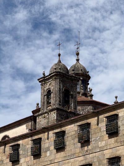 This is a close-up, vertical, low-angle shot of the exterior of an old stone building, likely a church or cathedral, in Santiago De Compostela, Spain. The building features ornate architectural elements, including domed towers topped with crosses, decorative finials, and arched windows. The facade is constructed from large, rough-hewn stone blocks. Several windows are visible, each protected by a black wrought-iron grate and containing small plants, suggesting they are decorative flower boxes. The sky above is partly cloudy with a bright, diffused light, indicating it is daytime. The image focuses on the intricate details of the building's stonework and religious symbolism, with no people or discernible activity visible. The weathered stone and the presence of moss and greenery growing in some crevices suggest the building has a significant historical presence. The perspective emphasizes the grandeur and age of the structure.