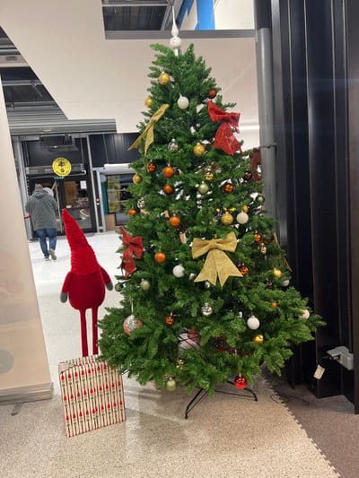 A decorated Christmas tree stands in an indoor space. It's adorned with gold, red, and orange ornaments, bows, and white spherical decorations. A red plush toy with long, thin legs and a striped present are positioned near the base of the tree. A person wearing a grey jacket is visible in the background. The area has a commercial or retail setting.