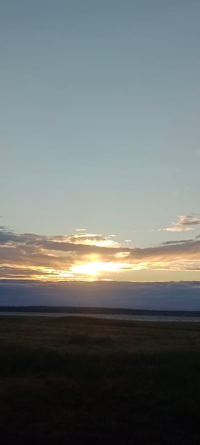 A vertical, full-frame shot captures a serene sunset over a body of water in Peskovatka, Russia. The sky, a soft gradient of pale blue and pale yellow, is adorned with scattered, wispy clouds. The sun, a brilliant orb, is partially obscured by a band of clouds, casting a warm, golden light that illuminates the edges of the clouds and creates a luminous glow on the horizon. Below the sky, a calm body of water stretches horizontally across the mid-ground. The foreground is dominated by a field of grass, depicted in dark, shadowy tones, suggesting the approach of twilight. The overall impression is one of tranquil natural beauty at the close of day.