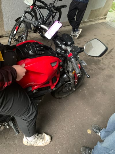 A red sport motorcycle is parked on a wet asphalt surface under a concrete overhang. A person wearing black pants and white athletic shoes is partially visible, sitting on the motorcycle and touching its red fairing. Another person in black pants and athletic shoes stands leaning against the concrete structure behind the motorcycle. In the background, a black bicycle is parked. The environment appears to be an outdoor urban space, possibly a parking area or alley in Severodvinsk, Russia.
