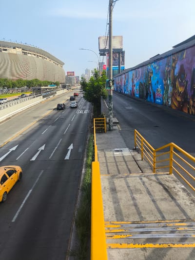 The photograph depicts a section of a highway in La Victoria, Peru. The image is taken from an elevated pedestrian walkway, with bright yellow railings visible in the foreground. To the left of the walkway is a multi-lane highway with multiple vehicles, including a yellow taxi. White directional arrows are painted on the road. On the left side of the highway, there's a large building, likely a stadium, under construction or renovation. To the right of the pedestrian walkway, a long wall is covered in colorful mural artwork. A pedestrian sidewalk is visible between the mural wall and the walkway. Billboards and other advertisements are displayed on poles and structures. The sky is clear and blue.