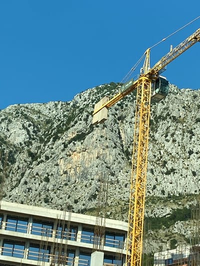 A yellow construction crane stands against a backdrop of a rugged, rocky mountainside with scattered vegetation. The crane's boom extends diagonally upwards and to the right, with a glass-enclosed operator's cab visible near the top of its vertical tower. In the foreground, a partially constructed building with multiple floors and dark-paneled windows is visible. The sky is a clear, bright blue. This scene is located in the city of Donji Katun, Montenegro.