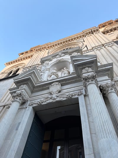 The image shows a low-angle view of the facade of the Basilica di San Sebastiano in Acireale, Italy. The facade is decorated with ornate Baroque sculptures and architectural details, including columns, reliefs, and statues of saints. A doorway is visible in the foreground, with a dark green door. The sky is clear and blue in the background.