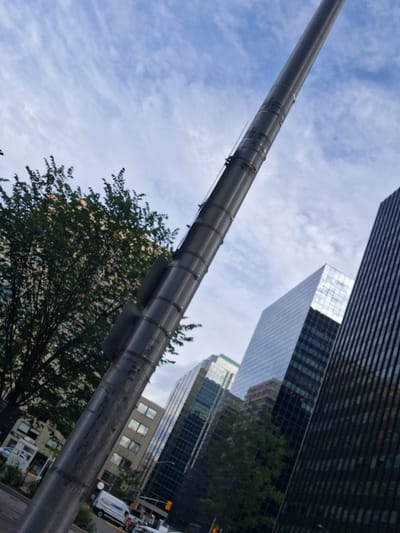 The image depicts a street scene with a large, cylindrical metal pole dominating the foreground, angled across the frame. In the background, several modern glass-fronted buildings are visible against a cloudy blue sky. A tree with dense green foliage is situated to the left of the pole. Below the pole and to the left, a white van with blue lettering and a smaller white kiosk-like structure are visible. Traffic lights and other street furniture are also present. The overall impression is an urban environment, likely in a business district, with a focus on architectural elements and infrastructure against a natural backdrop of trees and sky.