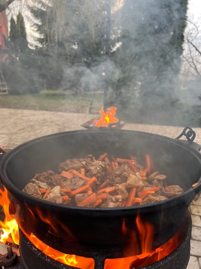 The image displays an outdoor cooking scene in Пролетарский, Russia, focused on a large, dark, cast-iron cauldron or kazan. This traditional cooking vessel is intensely heated by an open fire, with vibrant orange and yellow flames visible around its base and sides. Inside the cauldron, a dish composed of numerous chunks of meat and baton-cut carrots is being prepared. Significant plumes of white smoke billow from the fire and cooking process, spreading across the left side of the frame and partially obscuring the background. The cooking apparatus rests on paved ground, likely a patio or outdoor area. In the blurred background, the silhouettes of trees and foliage can be seen, indicating a natural outdoor setting. The overall impression is one of traditional outdoor cooking, possibly of a dish like pilaf or plov, common in Russian and Central Asian cuisine. No people or visible text are present in the image. The lighting suggests it is daytime, possibly late afternoon, given the atmospheric smoke.