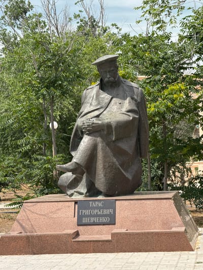 The image shows a bronze statue of a seated man in military attire. He is wearing a cap, a long coat, and boots. His hands are clasped in his lap, and he is looking down. The statue is mounted on a granite pedestal with a plaque that reads "ТАРАС ГРИГОРЬЕВИЧ ШЕВЧЕНКО" in Cyrillic. The statue is located in a park-like setting with trees and foliage in the background. The sky is visible above the trees. The ground in front of the pedestal is paved with light-colored bricks. The overall impression is of a monument to a historical figure, likely in a public space in Fort-Shevchenko, Kazakhstan.