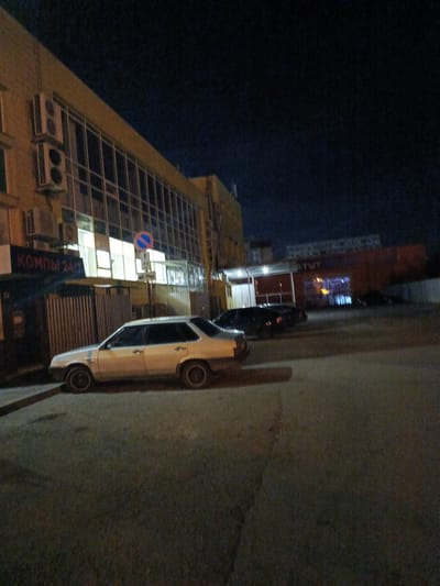 The image depicts a dimly lit street scene at night in Volzhsky, Russia. In the foreground, a light-colored sedan is parked on an asphalt surface. To the left, a section of a building facade is visible, featuring large windows and an air conditioning unit. A sign with Cyrillic text that reads "КОМПЫ 24/7" (COMPUTERS 24/7) is mounted on the building. Further down the street, another dark-colored vehicle is parked, and beyond that, another building with illuminated windows is visible. A traffic sign with a blue circle and a red diagonal line is also present. The overall environment suggests an urban commercial or residential area at night.