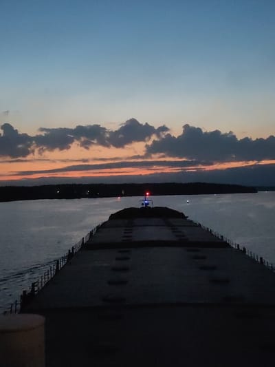 The image depicts a low-light scene from the deck of a ship, likely a freighter or bulk carrier. The ship appears to be traveling on a body of water, possibly a river or channel, as evidenced by the shoreline visible in the middle ground. The sky is layered with hues of blue, orange, and grey, indicating either sunrise or sunset. Clouds are scattered along the horizon. A few lights are visible on the shoreline and another one on the ship in the distance. The deck is dark and flat with some apparent manholes and railing along the side.
There is no information available in the image to determine if the location is in the city of Нова, Russia.