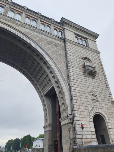 The image shows a large, stone archway with a tower on the right. The archway has decorative patterns and is supported by massive stone blocks. The tower is made of lighter colored stone blocks and has several windows. There is a small balcony on the tower. A traffic light with a red signal is visible in front of the tower. The sky is overcast. Trees and some buildings are visible in the background. The overall scene suggests an architectural structure of historical significance, possibly a bridge or part of a canal system. The location is Faliukovo, Russia.