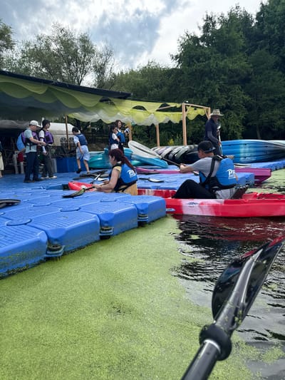 A group of people are gathered on a blue floating dock next to a body of water covered in green algae. Some individuals are wearing life vests and preparing to kayak, while others stand and observe. Several kayaks are visible, some on the dock and others in the water. In the background, trees and a cloudy sky are visible. The scene appears to be an outdoor recreational area. The location is not specified in the image itself, but this scene is consistent with various water sports locations in Lin'an District, China, which is known for its natural beauty and recreational activities.