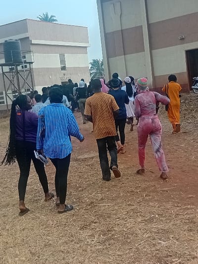 The image displays a group of approximately fifteen individuals walking away from the camera on a dry, unpaved reddish-brown path with sparse vegetation in Ayangba, Nigeria.
To the left, a multi-story building with alternating light and dark beige horizontal stripes is visible. A large black water tank on a metal stand is positioned in front of this building. A sign on its facade reads "TetFund." A palm tree is partially visible above and behind this structure.
To the right, a taller, multi-story building features alternating light and dark beige vertical stripes. A large, stylized emblem is present on its upper facade, and an open doorway is visible on its right side. A white motorbike is partially visible behind the doorway.
In the foreground, two females walk left-to-right. One, with long dark braided hair, wears a purple long-sleeved top and black leggings, with dark flip-flops, looking to her left and smiling. Adjacent, a female wears a blue and white vertically striped long-sleeved shirt, black leggings, and dark sandals, holding white papers in her left hand.
Further into the scene, a male wearing a brown and yellow striped short-sleeved shirt and dark pants walks away. To his right, a person in a pink, red, and white tie-dye full-body garment and a matching head covering moves in the same direction. The remaining individuals in the mid-ground and background are dressed in various casual attire, including dark, light, and an orange full-body garment on the far right. The overall lighting is diffused.