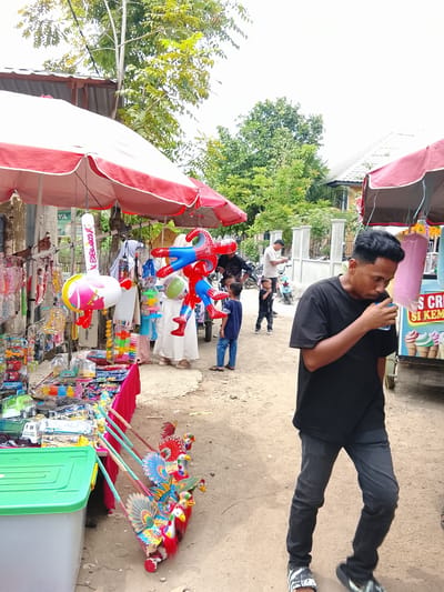 A street scene in Rawasari, Indonesia features a stall selling toys under a red and white striped umbrella. Several children are present, including two boys in the foreground wearing blue and black shirts respectively, and another boy in a black t-shirt and pants further back. A man in a black t-shirt and dark jeans is walking past, drinking from a cup. Behind the stall, people in white clothing are visible, and a pink cotton candy machine is on display. The ground is dirt, and trees and buildings are in the background.