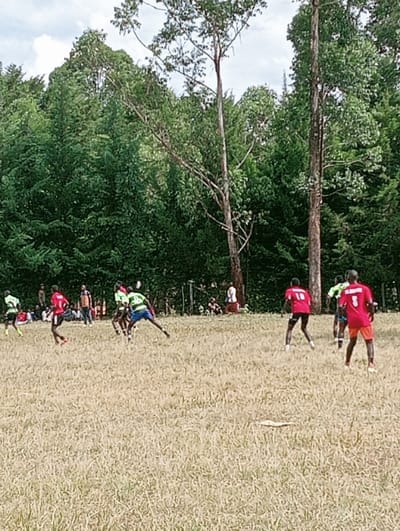 This image depicts an outdoor daytime scene in Lwandeti ward, Kenya, featuring a sporting event on a field with sparse, dry, light brown, and yellowed grass. Approximately 10 individuals are actively engaged in play across the midground and foreground. Five players wear red jerseys, one on the far right displaying the number '3' on his back, and another in the mid-right displaying the number '10'. Three players wear green and white striped jerseys, one in the mid-left captured in a leaning, active posture. Several other individuals are visible in the background, gathered beneath a dense line of dark green trees. Two significantly taller, slender trees with lighter bark and sparser foliage rise above the main treeline against an overcast sky featuring white and grey clouds.
