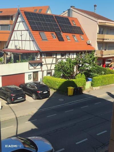 This media file depicts a sunny day in Constance, Germany. The scene shows a traditional German half-timbered house with a red-tiled roof and solar panels installed on the upper section of the roof. Next to it is a light pink building with balconies. In front of the half-timbered house, there is a red garage door, and two black SUVs are parked on a driveway. A neatly trimmed green hedge runs along the front of the property, and beyond that, a street with a blue recycling bin and several trash cans, including a yellow one, are visible. Lush green trees and bushes are also present. The time of day appears to be late morning or early afternoon, judging by the bright sunlight and the shadows cast by the buildings and trees. The clear blue sky indicates fair weather. The presence of the traditional architecture, the German license plates on the cars (KN-TL 23 and KN-MH 399E), and the city name "Constance" in the provided context pinpoint the location to Constance, Germany. The image is watermarked with "XIAOMI 12" in the lower left corner. There is no activity or event taking place; it is a static scene of a residential area.