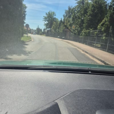 The image is taken from the interior of a vehicle, looking forward through a dirty windshield. The view is of a road in the city of Bralin, Poland, on a sunny day. The road is asphalt and appears to be a two-lane street with a paved shoulder and a sidewalk on the right side. Trees line the right side of the road, and some buildings and a fence are visible in the background. There are some vehicles on the road in the distance. The foreground of the image shows the dashboard of the car, which is dark gray and textured. The scene depicts a typical view of driving through a town.