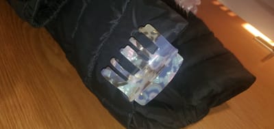 This media file shows a close-up shot of a translucent, pearlescent hair comb resting on a dark, puffy fabric, likely a jacket or cushion. The comb has a unique, angular design with several teeth. The fabric it lies on has a quilted texture with visible stitching. The background consists of a light brown wooden surface, possibly a floor or table, with a bright, angled light source casting elongated shadows. The lighting suggests it is daytime, with a strong artificial or natural light source. No people are visible in the media file. The scene appears to be indoors, given the presence of a wooden surface and controlled lighting. The overall aesthetic is simple and focuses on the textures and colors of the object and its immediate surroundings.