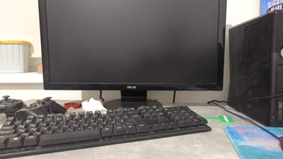 A close-up, eye-level shot captures a desk setup, likely in a home environment in Zadebri, Ukraine. Dominating the frame is a large, black computer monitor, its screen dark and reflecting the room. Below the monitor rests a black keyboard, its keys showing dust accumulation, suggesting it's well-used. To the left of the keyboard are two black video game controllers, positioned side-by-side. To the right of the monitor, a black Xbox 360 console is visible, its casing also appearing dusty. A collection of miscellaneous items are scattered across the desk, including a white object, possibly a dust cover or figurine, and a red, circular object with a woven pattern, perhaps a coaster or bracelet. In the background, to the left of the monitor, a white and yellow storage container sits on a shelf, and a small spool of thread or wire is visible behind it. The overall scene is one of a functional, though slightly cluttered, gaming or computer station.