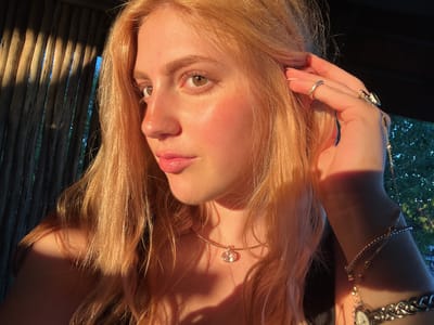 A close-up, upside-down portrait of a woman with light blonde hair, hazel eyes, and fair skin, bathed in warm light. She is wearing a delicate gold necklace with a gemstone pendant, a thick chain bracelet with a watch, and multiple rings on her left hand, which is near her ear. The background features vertical wooden slats, partially illuminated by sunlight. The location is Khwai, Botswana.