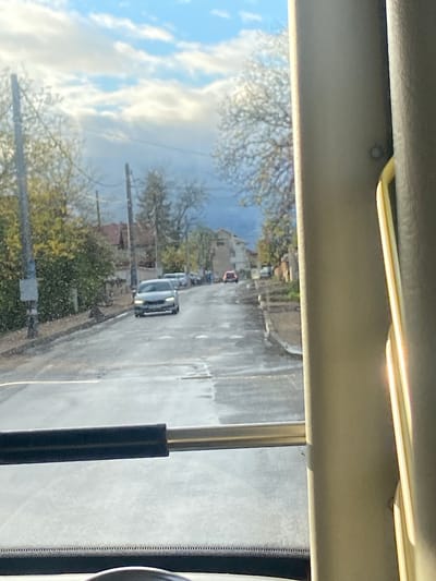 The image captures a daytime street scene from the perspective of a passenger inside a vehicle, looking out a side window. The view is partially obscured by the vehicle's interior elements.
In the foreground, the top of the image shows the light-colored ceiling or headliner of the vehicle, along with a sun visor and a yellow-white plastic trim piece. Towards the right side of the frame, a black and silver pillar, likely part of the window frame or a grab handle, is visible.
Outside the vehicle, a residential street in Golyanovtsi, Bulgaria, is visible under a partially cloudy sky. The road surface appears wet and mottled, suggesting recent rain or puddles. A light-colored car, possibly white or silver, is parked or slowly moving on the street, facing away from the viewer. Further down the street, a red car is also discernible.
Along the left side of the street, there are several trees, mostly with bare branches, though some show hints of early spring foliage. Utility poles run alongside the road. Beyond the trees, the bright blue sky with scattered white clouds is prominent. On the right side of the street, more trees with green foliage and some distant buildings, likely houses, can be seen. No discernible text is visible in the image.