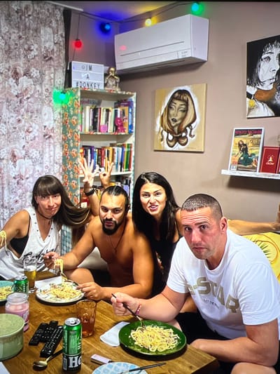 Four individuals, two men and two women, are seated around a wooden table, engaged in eating pasta. The environment appears to be an indoor setting, possibly a dining area within a residence. The wall behind them is adorned with various decorative items, including paintings of faces, a shelf filled with books, and string lights. An air conditioning unit is visible mounted on the wall above. On the table, plates of spaghetti, drinks in glasses and cans, remote controls, and other miscellaneous items are present. The lighting in the room is a mix of overhead and string lights, creating a somewhat casual and eclectic atmosphere. The people are dressed casually, and their expressions suggest a relaxed, social gathering. It is important to note that there is no indication or information within the image to suggest the location is in the city of Mollet del Vallès, Spain.