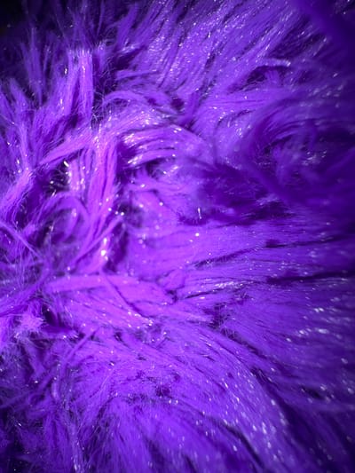 The image shows a close-up of purple faux fur with silver tinsel threads woven throughout. The fur is arranged in a swirling pattern, and the lighting creates a soft, diffused effect with bright highlights on the tinsel. No location or people are visible.