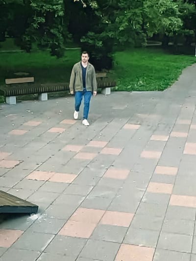 A man walks on a paved area in front of a park. The man is wearing a green jacket over a dark t-shirt and blue jeans, with white sneakers. The paved area consists of square tiles in shades of grey and peach. In the background, there is a green lawn with trees and park benches. The scene appears to be in Dondușeni, Moldova, given the context.
