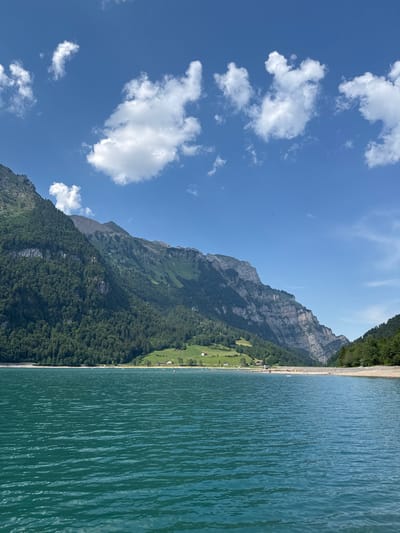 This is a landscape photograph of Walenstadtsee in the canton of St. Gallen, Switzerland, not Glarus. The image captures a bright, sunny day with a clear blue sky dotted with white, fluffy clouds. In the foreground, the turquoise water of the lake is rippled by a gentle breeze. The middle ground features a pebble beach with several people visible, some appearing to be sunbathing or wading in the water. A few small boats are also present on the lake. In the background, lush green mountains rise steeply, covered in dense forest. The rocky faces of the mountains are exposed in some areas, showcasing their rugged beauty. A few scattered houses and chalets are nestled on the lower slopes of the mountains, adding a touch of human habitation to the natural scenery. The overall impression is one of a serene and picturesque alpine lake environment on a beautiful summer day.
